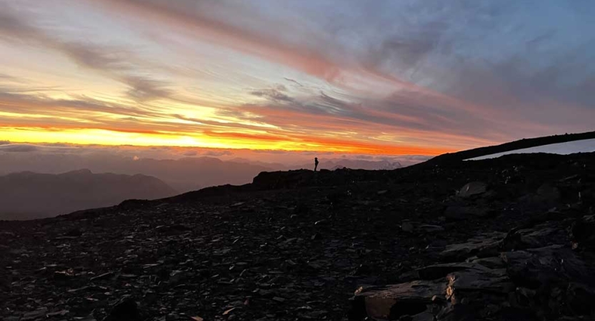 From a distance, the silhouette of a person standing on an overlook is illuminated against a yellow, orange, purple and blue sky. 